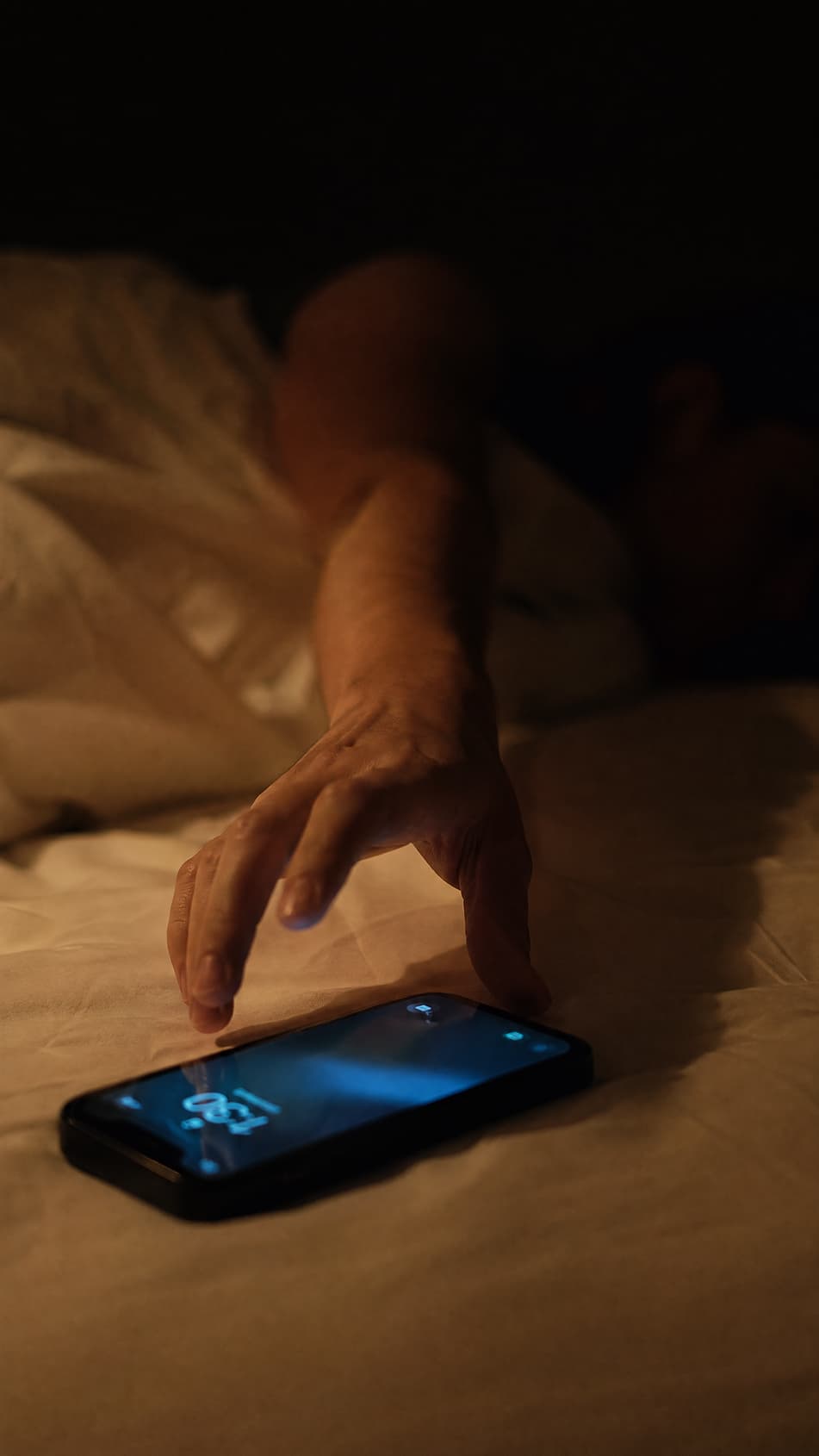 A hand reaching for a phone on a bed in low light